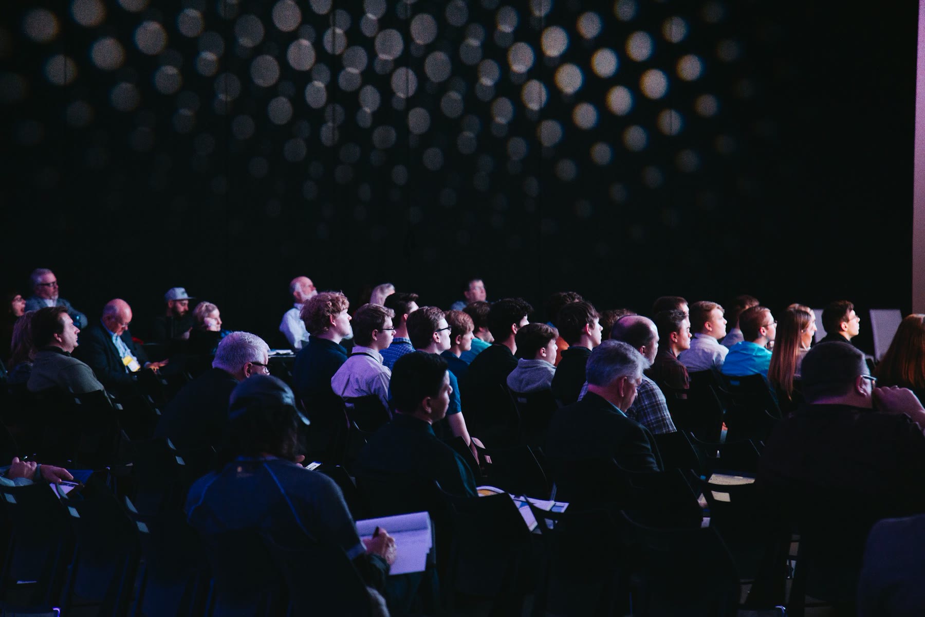 A packed conference audience seated in rows, facing a stage and large presentation screens.