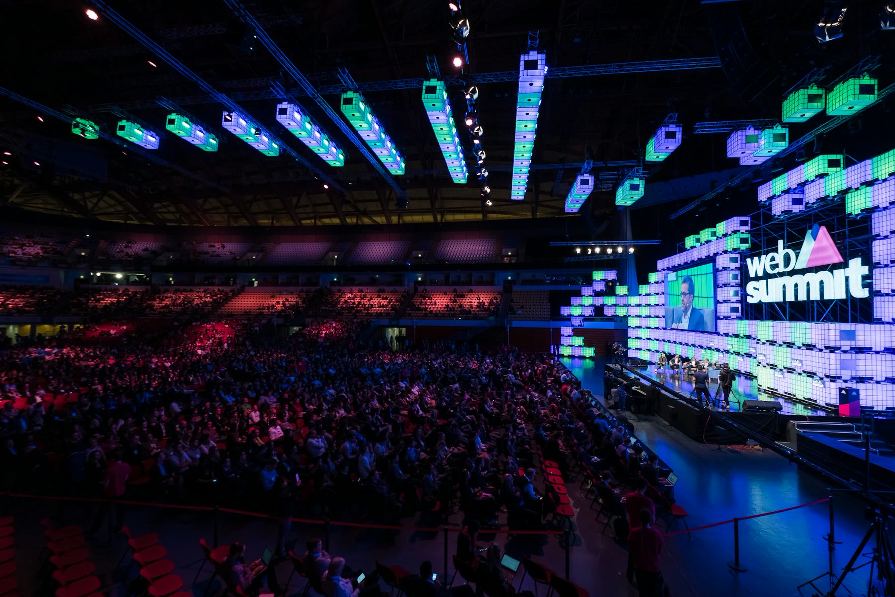 A crowded tech conference floor, with people walking between exhibition booths and large screens.